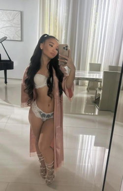 A woman with long dark hair takes a mirror selfie in white lace lingerie and a pink robe, standing in a bright, modern