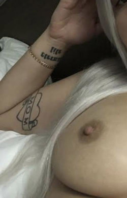 Blonde woman with white hair, visible tattoo on arm and wrist, lying down with bare chest partially exposed.
