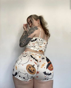 Blonde woman with tattoos in Halloween-themed crop top and shorts, posing sideways against a plain wall.