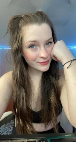 A young woman with long brown hair and blue eyes rests her head on her hand, wearing a black top in a room with blue