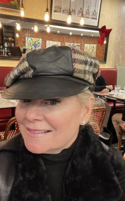 A woman with blonde hair wearing a black and plaid cap, black turtleneck, and fur-trimmed coat, smiling in a restaurant