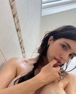 Brunette woman with wet hair in a bathroom, topless, touching her lips, taking a selfie.