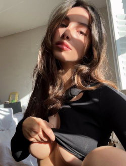 Brunette woman with long hair in a black top, sitting indoors near a window, pulling fabric to reveal cleavage.
