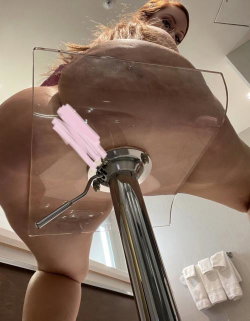 A woman with a curvy build bends over in a bathroom, her backside visible near a chrome shower fixture; she wears a