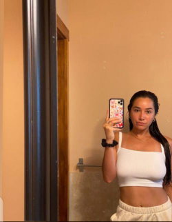 A woman with long dark hair takes a mirror selfie in a white crop top and light-colored pants, standing in a bathroom