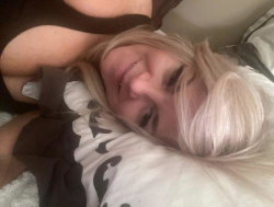 Blonde woman with a curvy build lying on a bed, wearing a dark top, resting her head on a pillow.