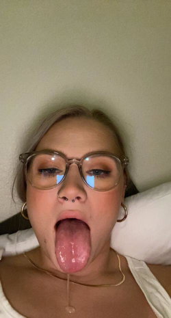 A woman with blonde hair and glasses lies on a bed, sticking her tongue out while wearing a white top and gold jewelry.