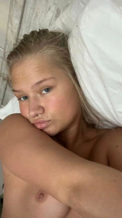 Blonde woman with nose ring lying in bed, partially covered by white sheets, bare chest visible.