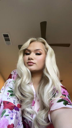 Blonde woman with long wavy hair wearing a floral robe, taking a selfie indoors under a ceiling fan.