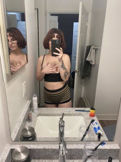 A woman with short brown hair takes a mirror selfie in a bathroom, wearing a black crop top and striped shorts, hand