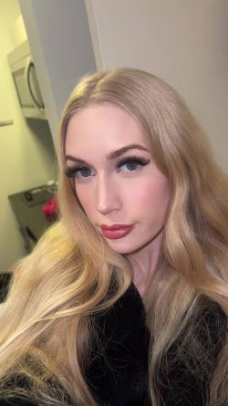 Blonde woman with long wavy hair and full makeup, wearing a black top, taking a close-up selfie indoors.