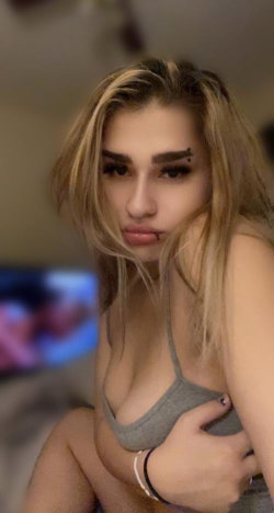 A woman with blonde hair and a nose piercing wears a gray tank top, posing indoors with one hand on her chest.