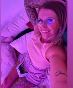 A smiling woman with blonde hair and round glasses lies on a bed in a pink-lit room, wearing a sleeveless top and