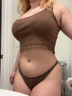 Curvy woman with blonde hair wearing a brown ribbed crop top and matching thong, standing in a bathroom.