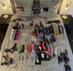 Various sex toys and adult products arranged neatly on a bed with gray sheets, between two bedside lamps.