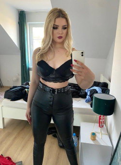 Blonde woman in black leather-like crop top and pants takes a mirror selfie in a bedroom with white furniture and green