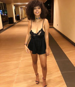 A woman with curly hair and a black lace dress stands in a hallway.