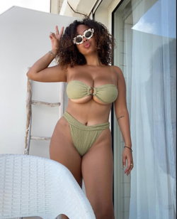 Curvy woman in a green bikini posing by a window with sunglasses.