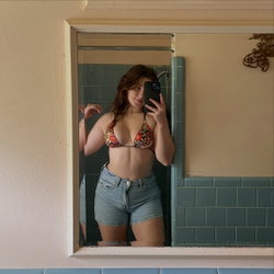Curvy woman with reddish-brown hair in floral bikini top and denim shorts takes a bathroom mirror selfie.