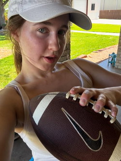 A young woman in a white cap and tank top holds a football outdoors on a sunny day, looking at the camera.