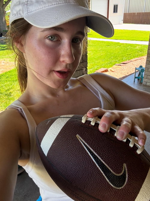 A young woman in a white cap and tank top holds a football outdoors on a sunny day, looking at the camera.