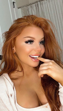 Curvy redhead smiles playfully with finger on lips in bathroom, wearing open white cardigan.