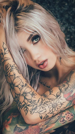 Blonde woman with extensive tattoos on her arms rests on a dark textured surface.