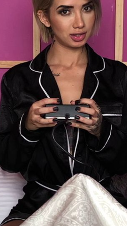 Woman with short blonde hair in black pajamas holding a video game controller against a pink wall.