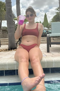 Woman in red bikini sits poolside holding a drink with palm trees in the background.
