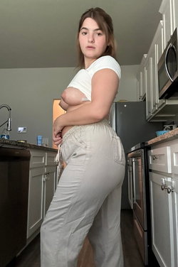 A curvy brunette stands in a kitchen wearing a white top and light pants.