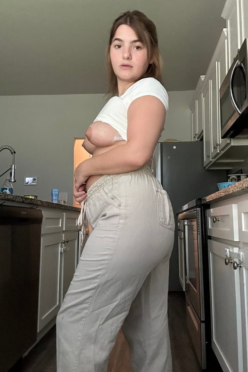 A curvy brunette stands in a kitchen wearing a white top and light pants.