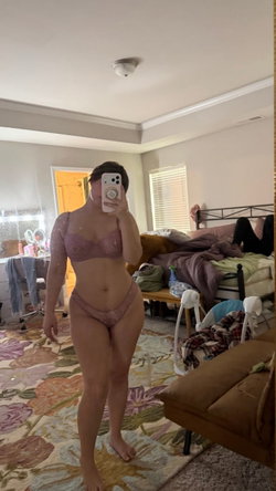 A curvy brunette woman wearing pink lace lingerie takes a mirror selfie in her bedroom.