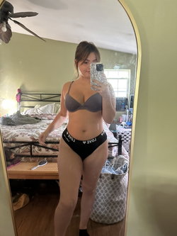 Curvy woman in gray bra and black thong takes mirror selfie in bedroom, holding phone with patterned case.