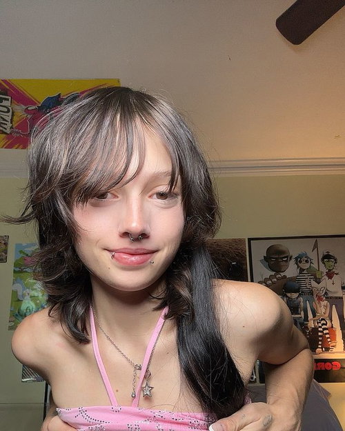 Slim woman with shaggy dark hair, pink spaghetti strap top, in bedroom with posters.