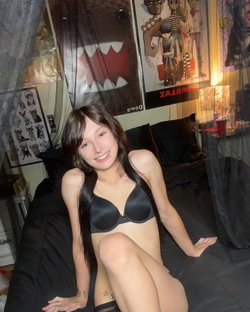 Slim brunette in black bra smiles on bed in dimly lit room with posters.