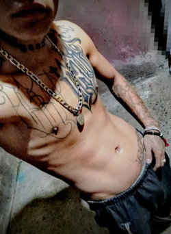 Shirtless muscular man with extensive chest and arm tattoos, wearing a chain necklace and dark shorts, standing indoors.