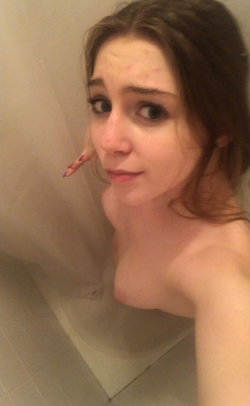 A young woman with brown hair takes a close-up selfie in a bathroom, bare-chested and looking directly at the camera.