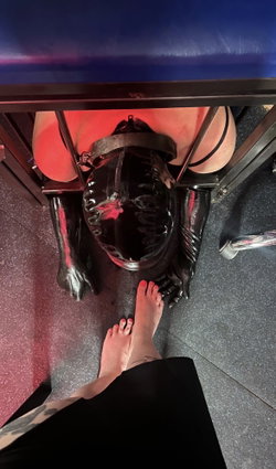 A person in a shiny black latex hood and gloves is restrained under a table, viewed from above; bare feet with red