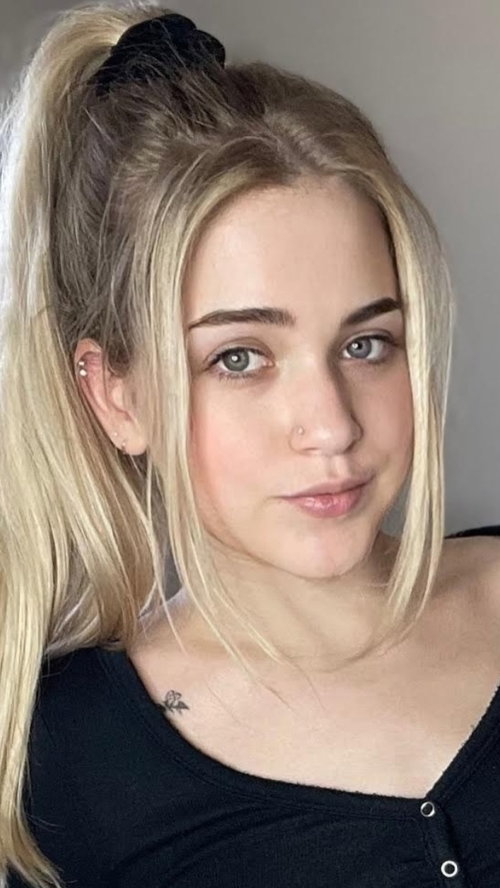 Blonde woman with a high ponytail and nose ring, wearing a black top, looking directly at the camera in a close-up
