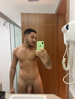 A shirtless man with tattoos takes a selfie in a bathroom.