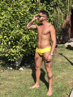 A shirtless man in yellow swim trunks stands in a garden, adjusting his sunglasses.