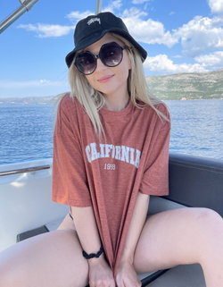 Blonde woman wearing a bucket hat and sunglasses sits on a boat in a California t-shirt.
