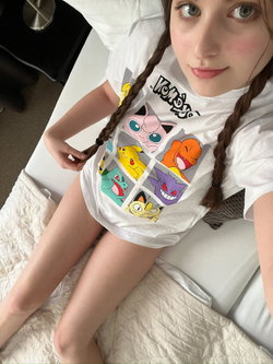 A young woman with braided brown hair lies on a bed, wearing a white graphic t-shirt and no visible bottoms, looking at