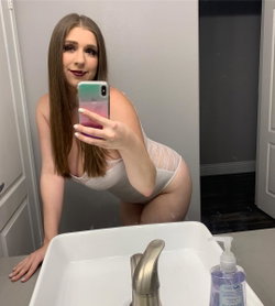 Brunette woman with curvy figure takes mirror selfie in white lace bodysuit, leaning over bathroom sink.