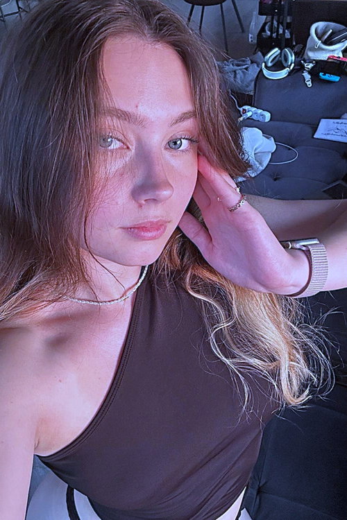 Slim woman with wavy brunette hair and blonde tips wears one-shoulder brown top in messy room.
