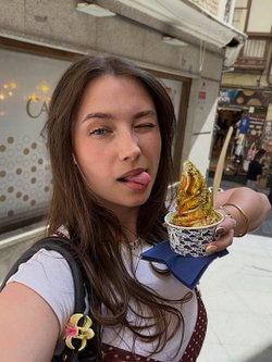 Brunette woman with long hair winks and sticks out her tongue while holding a dessert cup in a city street, wearing a