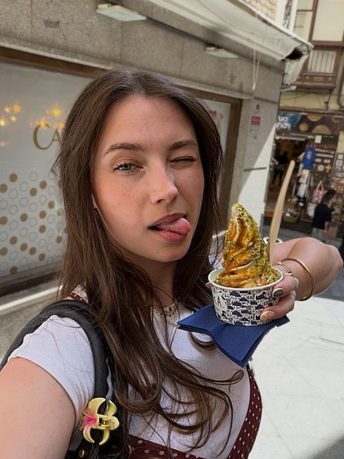 Brunette woman with long hair winks and sticks out her tongue while holding a dessert cup in a city street, wearing a