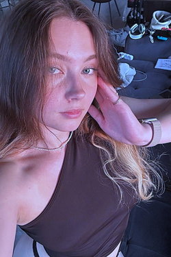 Young woman with long brown hair takes a close-up selfie indoors wearing a brown tank top.