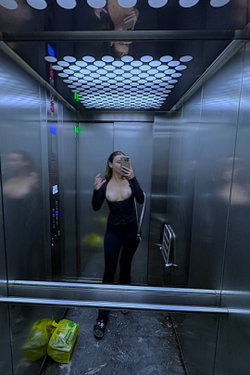 A woman in a black outfit takes a selfie in an elevator with a patterned ceiling.