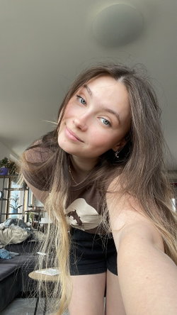 Smiling woman with long wavy brown hair wearing brown bear-print top and black shorts, indoor selfie.